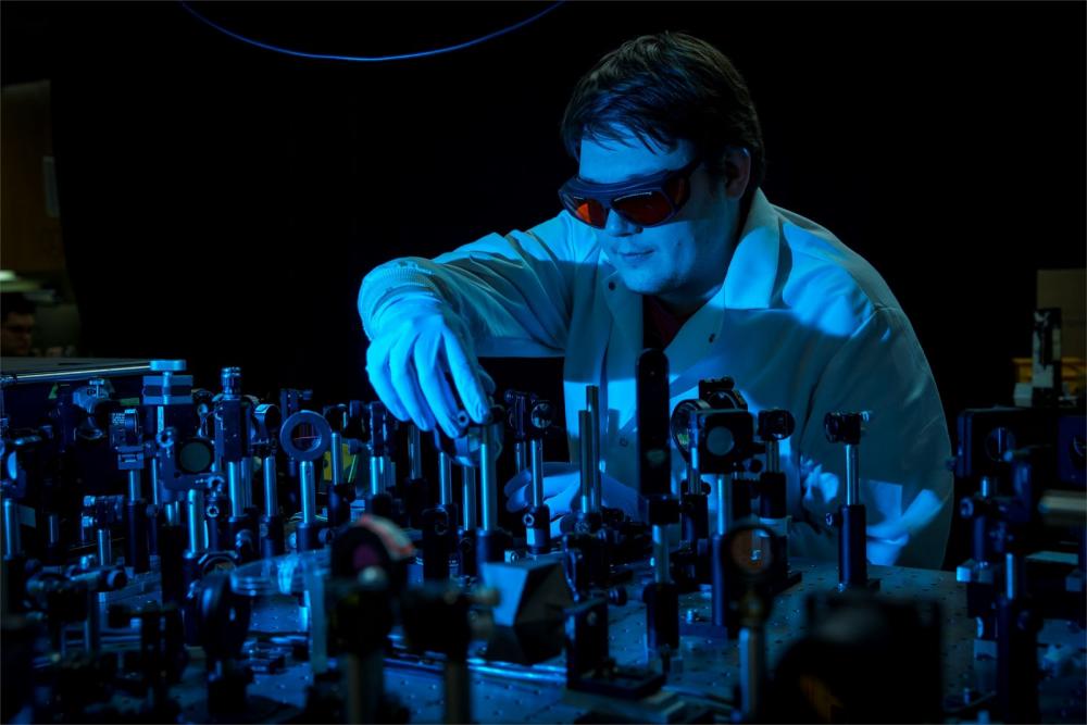 Temple chemistry professor wearing a labcoat, safety goggles and gloves in a darkened lab touching a device amidst many similar devices.