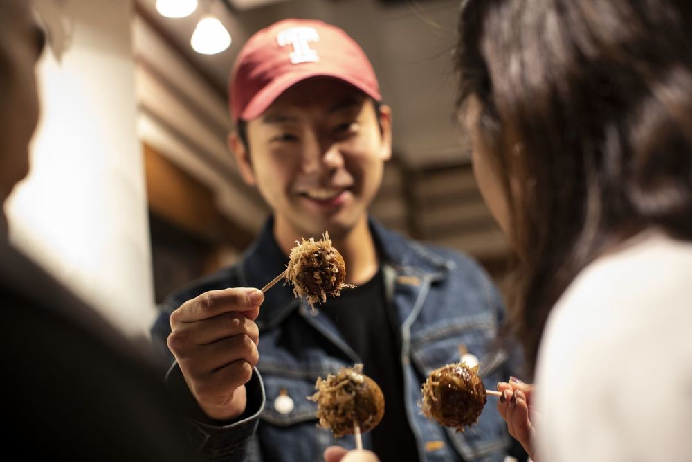 A smiling student wearing a red Temple University hat holds up a dumpling on a toothpick.