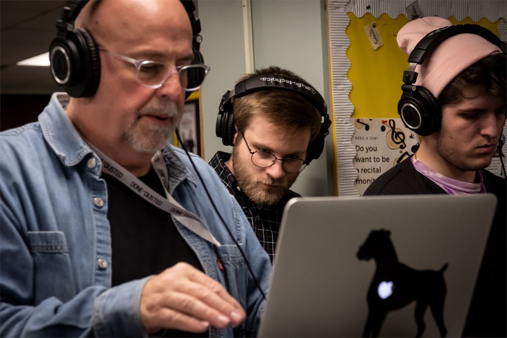 A professor with headphones works on a computer while two students also wearing headphones work alongside him.